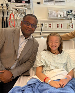 The transplant patient, Mary, in a hospital bed wearing a medical gown and covered with a blue blanket next to Dr. Andre Dick. The room includes medical equipment, monitors, and supply panels on the wall.