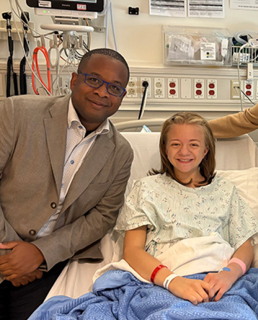 The transplant patient, Mary, in a hospital bed wearing a medical gown and covered with a blue blanket next to Dr. Andre Dick. The room includes medical equipment, monitors, and supply panels on the wall.