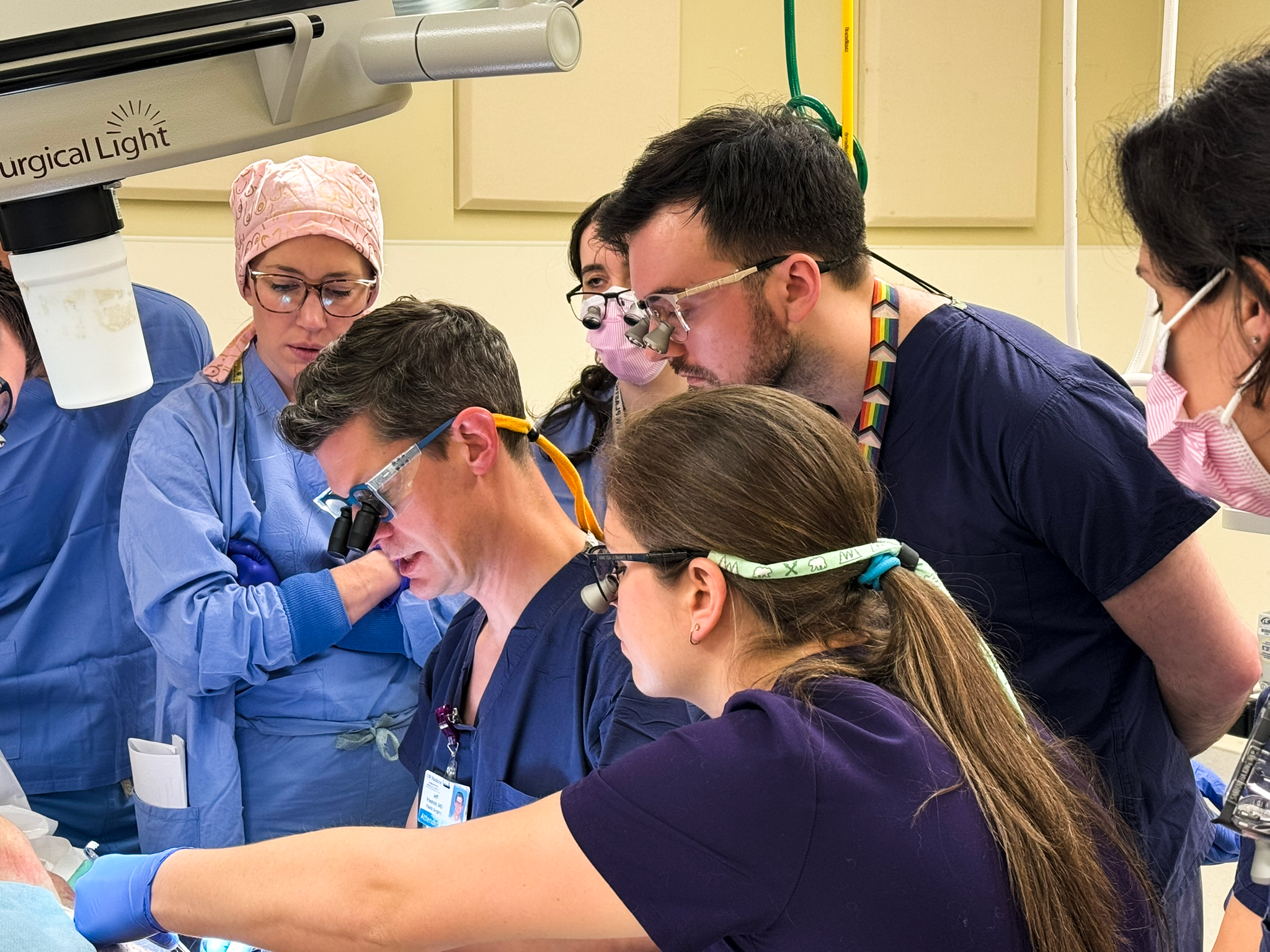 A multidisciplinary surgical team in scrubs collaborates around an operating table under a surgical light during a procedure or training scenario.