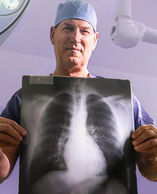 Dr. Douglas Wood, The Henry Harkins Professor & Chair, wearing surgical scrubs and a cap holds a chest X-ray showing the lungs. Overhead surgical lights and equipment are visible in the background.