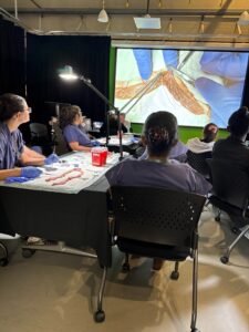 Participants in a surgical skills laboratory practice vascular anastomosis techniques at workstations while an instructor's demonstration is displayed on a projection screen.