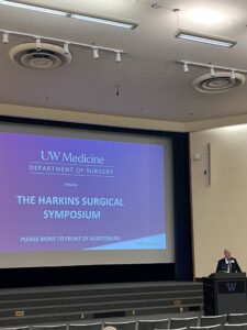 Presentation slide for the UW Medicine Department of Surgery Harkins Surgical Symposium displayed on an auditorium screen, with a speaker at a podium.